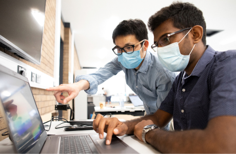 Two men wearing surgical masks and looking at information on a laptop computer