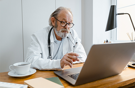 A doctor at his desk wearing a white coat and stethoscope looking at a computer 