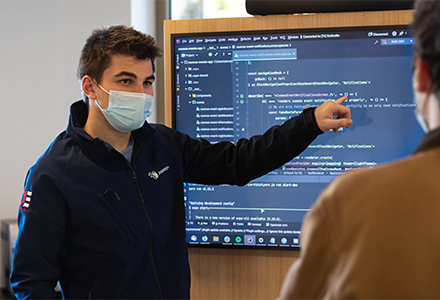 Person in a mask pointing at a computer screen and talking to another person