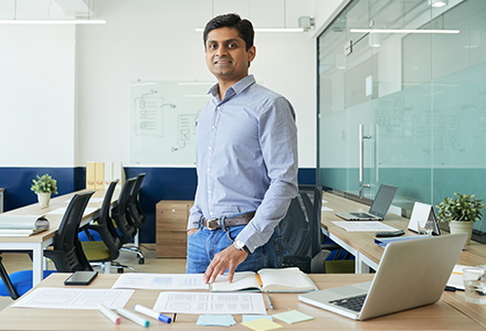 Man standing in a confereance room, he is smiling and wearing a blue shirt and jeans.