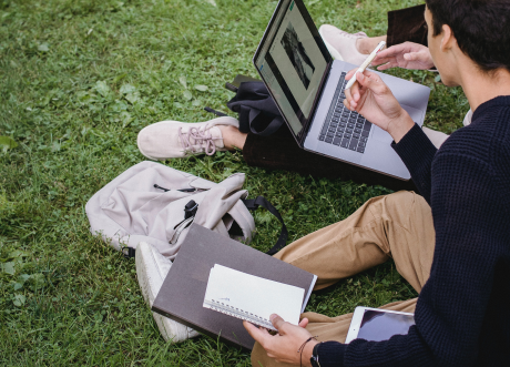 Students sitting on a grass looking over a laptop and taking notes on a notebook.